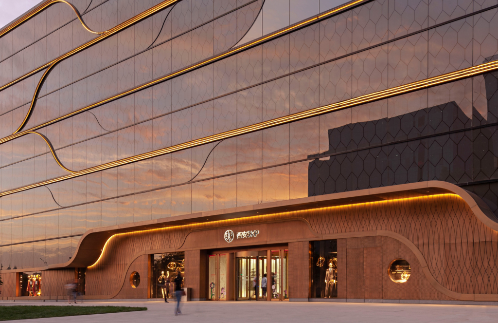 The SKP Xi’an façade expresses the SKP Curve at urban scale, with illuminated bronze-toned lines, reflective glazing and a flowing entrance canopy creating a distinctive luxury retail frontage at dusk.