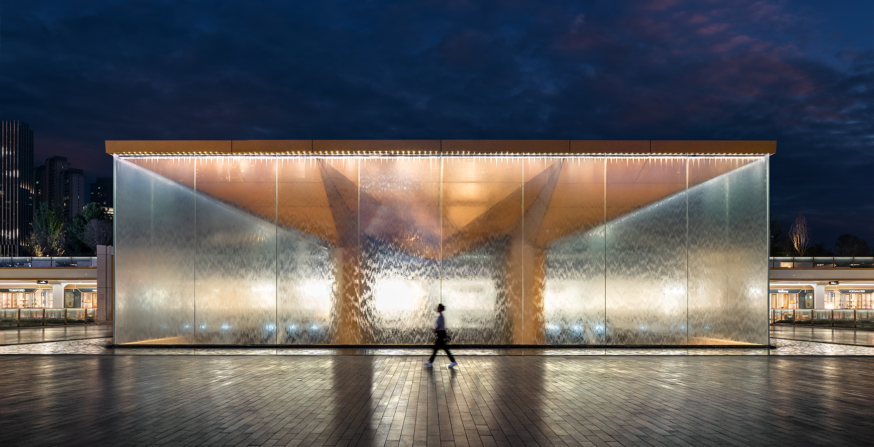 Illuminated glass pavilion at SKP Chengdu at night, with water cascading over the façade and a pedestrian passing in front.