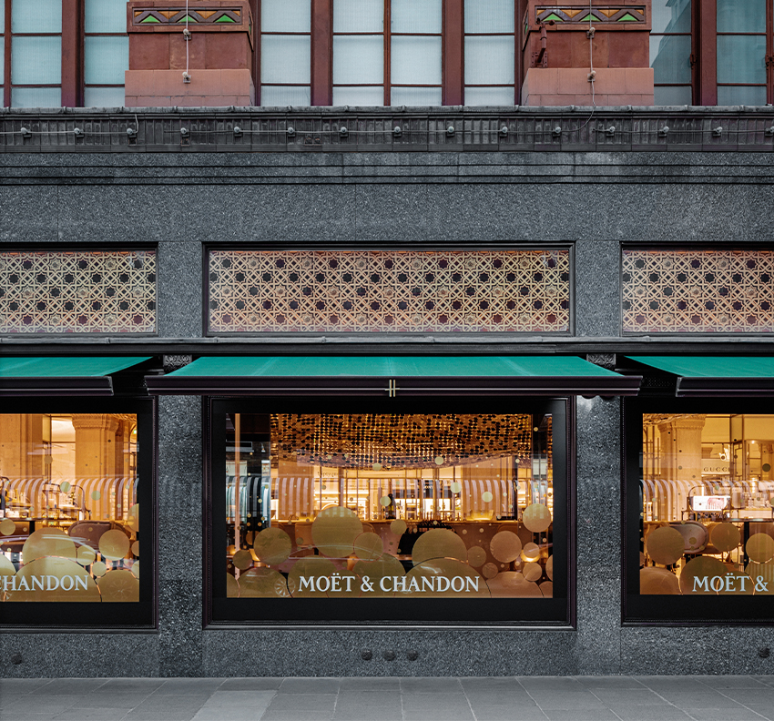 The Moët & Chandon Champagne Bar at Harrods presents a richly layered façade, with patterned screens, deep green awnings and warm interior lighting creating a refined hospitality presence within the London department store.