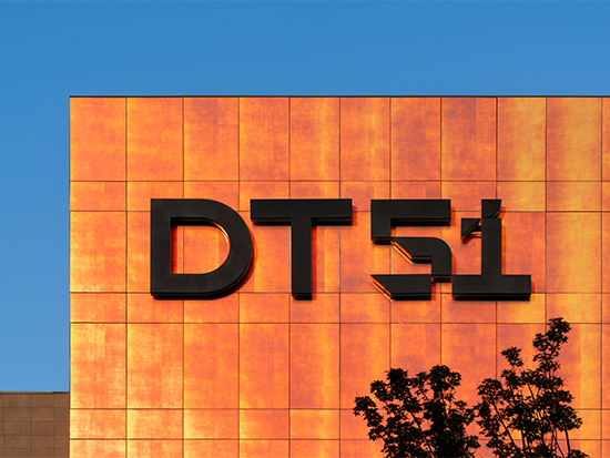 DT51 presents a bold architectural identity through its illuminated façade, where warm metallic cladding and strong graphic signage create a distinctive retail landmark against the city skyline.