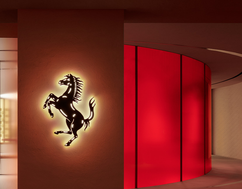 The Ferrari retail environment frames the brand’s emblem within a warm architectural façade, using terracotta tones, glazed transparency and sculptural interior details to create a refined expression of performance-led luxury.
