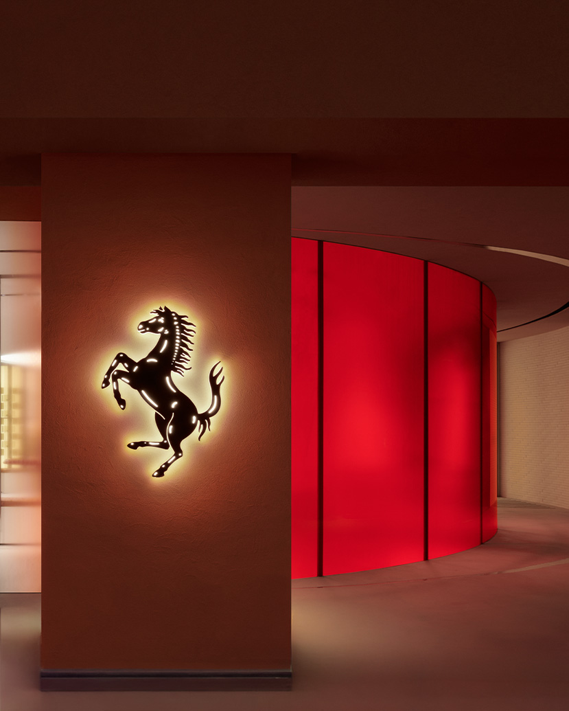 The Ferrari retail environment frames the brand’s emblem within a warm architectural façade, using terracotta tones, glazed transparency and sculptural interior details to create a refined expression of performance-led luxury.